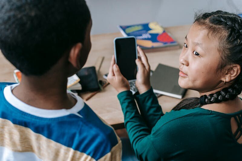 From above of adorable little Asian girl showing mobile phone to unrecognizable African American boy during break in classroom