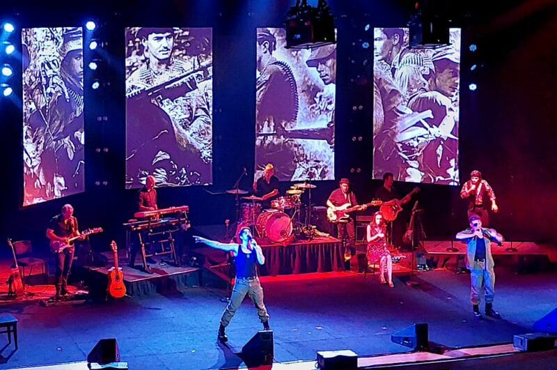 Hudobný koncert kapely s obrazmi vojakov na veľkých obrazovkách.