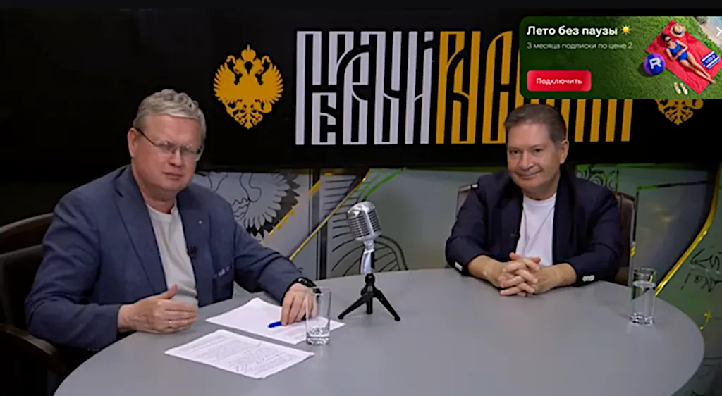 Dvaja moderátori sedia za štúdiovým stolom s mikrofónom.